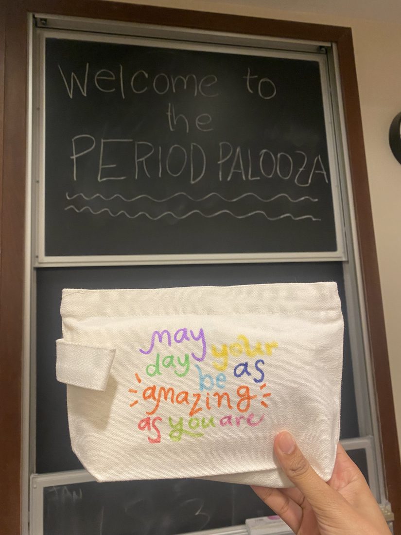 A canvas pouch that says 'may your day be as amazing as you are' in colorful marker being held up in front of a chalkboard that says 'welcome to the period palooza' on it.