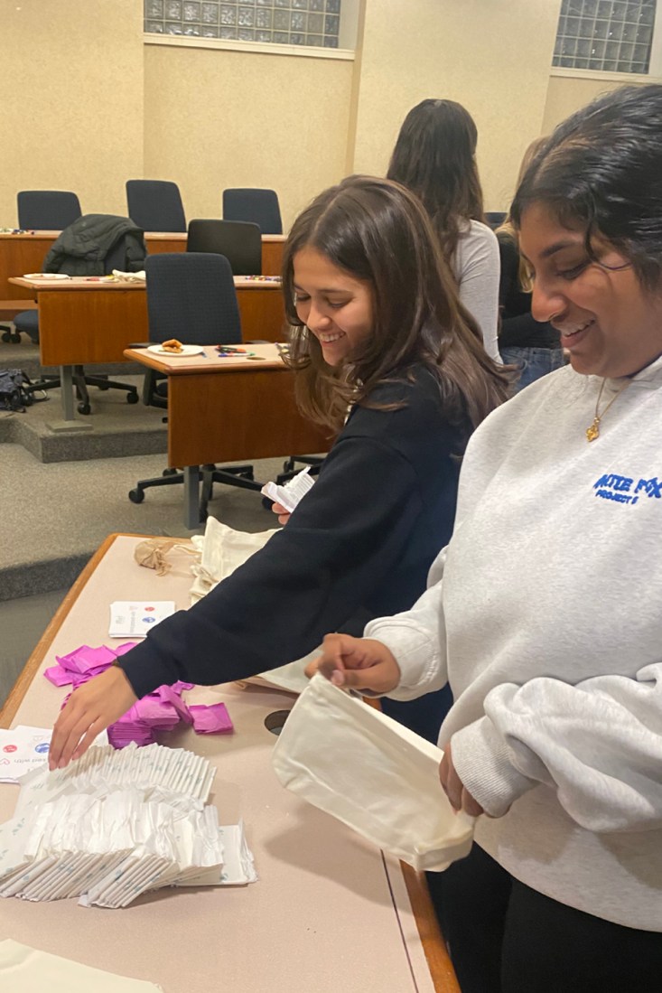 Two people standing at a table full of menstrual products filling canvas pouches with them.