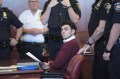 Luigi Mangione wearing a maroon sweater over a white shirt with a collar sitting in court holding a sheet of paper. Around him are several police officers in uniform.