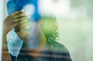 A female physician in scrubs and a surgical cap rests her forehead on her hand, appearing exhausted.