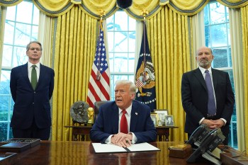 President Donald Trunp speaking while sitting in the Oval Office. Secretary Scott Bessent and Howard Lutnick stand on either side of him listening.