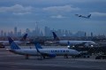 Two airplanes on a runway. In the background, a third airplane is taking off in front of a city skyline.