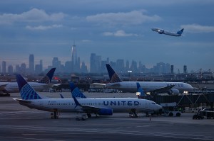 Two airplanes on a runway. In the background, a third airplane is taking off in front of a city skyline.