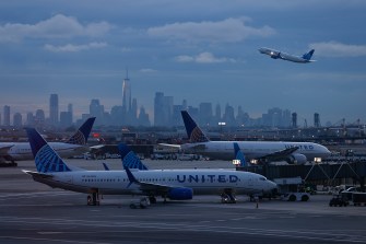 Two airplanes on a runway. In the background, a third airplane is taking off in front of a city skyline.