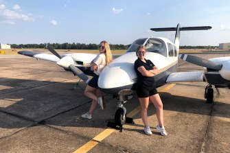 Calli Colvin and another person both posing with their backs up against a small plane on a runway.