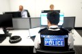 A person wearing a police branded vest sitting at a desk with a laptop and monitor in front of them. On the other side of the desk, two other people sit in front of computers.