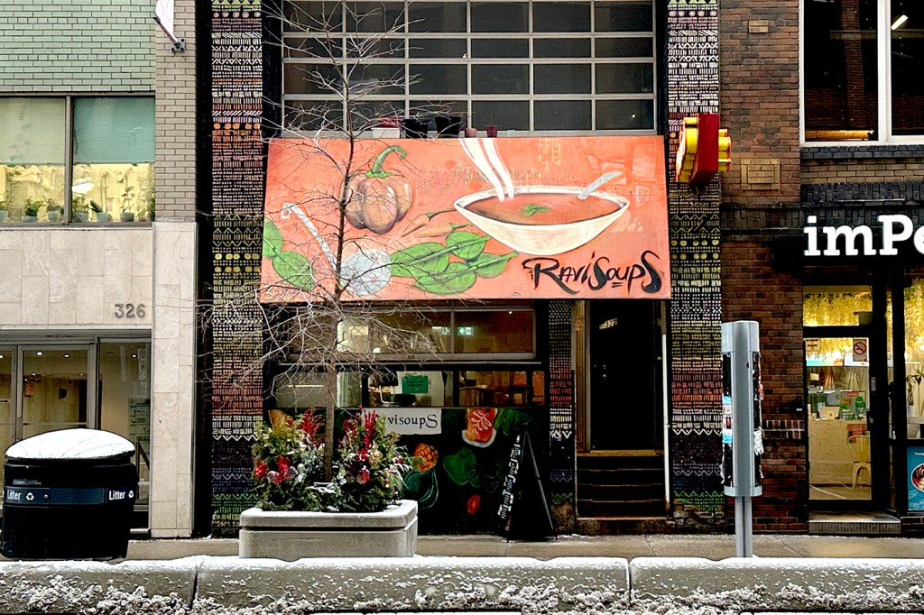 A shop on a street in Toronto with a reddish orange overhang that has a painting of a bowl of soup and the restaurant name 'Ravi Soups' on it.