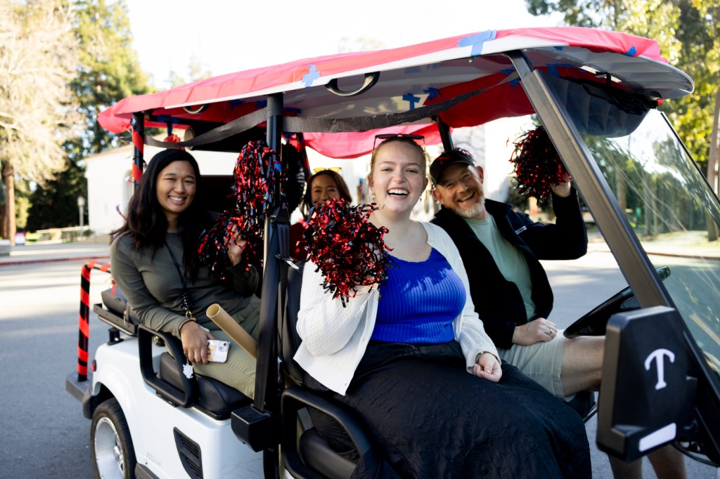 A group of people riding in a decorated golf cart, smiling and waving pom-poms.