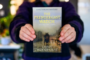 A person holding up a copy of The Prendergast Watch. The cover has the title on it at the top in yellow all caps font. At the bottom is Emma Woodhouse' name in all caps white text. A victorian coach on a road is depicted at the center of the cover.