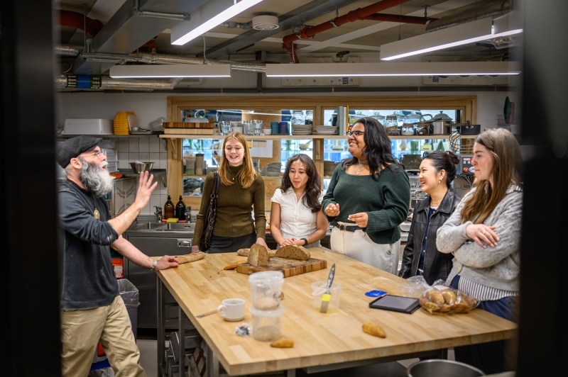 Five women in casual clothing stand around a wooden table with sliced bread and a cutting board as a bearded man in a black skullcap gestures while speaking to them from across the table.