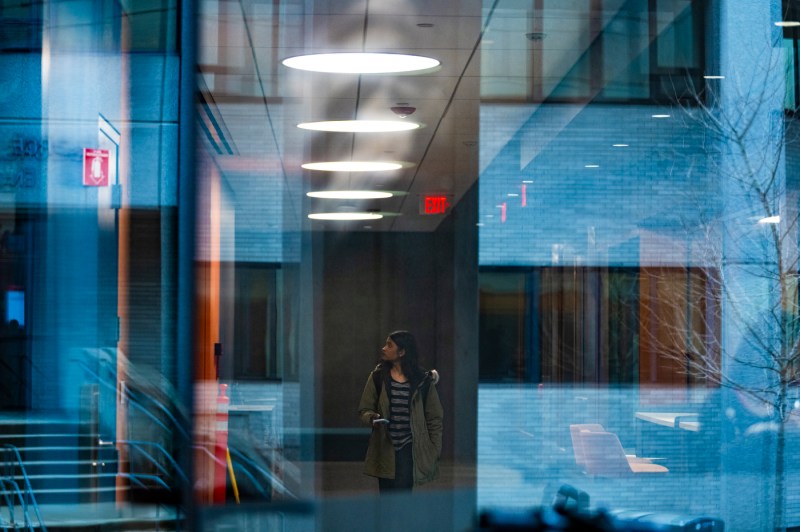 A person walks through an illuminated indoor space, seen through layers of glass and reflections.