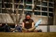 A student sits on the grass outdoors, working on a laptop with headphones on, enjoying the sunny weather on campus.