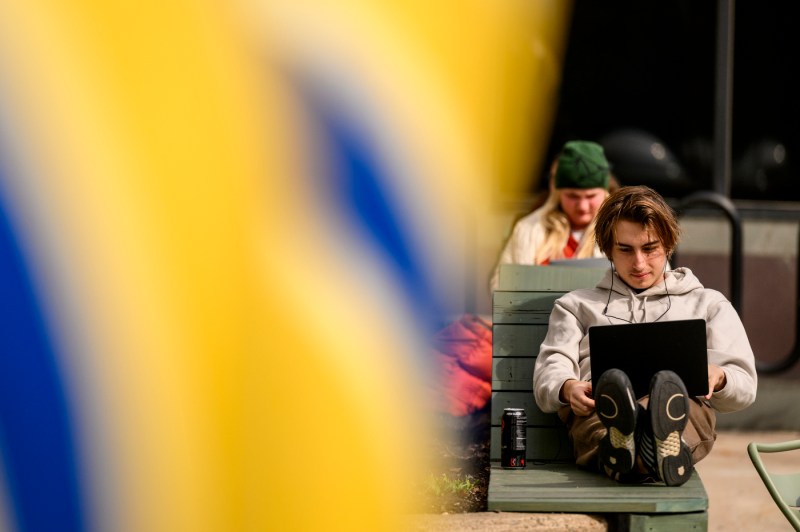 A person sitting outside with a laptop, appearing focused, while another person sits nearby. A blurred yellow and blue object is in the foreground.