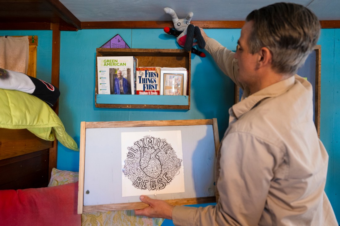 Alex Eaves holding a print in one hand and resting his hand against the wall of his U-Haul truck home in the other. There is a shelf of books on the wall next to him.