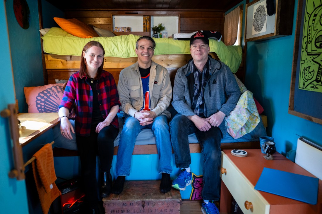 Alex Eaves, Derek Diedricksen and Amy Henion sitting on a twin sized bed inside of a box truck tiny house. 