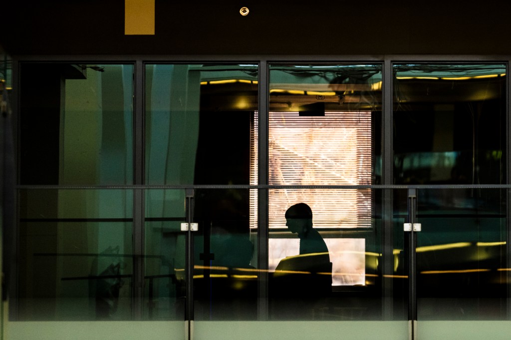 A silhouette of a person is visible through windows in an indoor setting, with reflections and shadows creating an abstract scene.