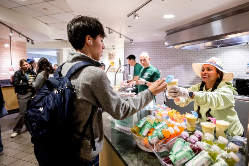 A person hands a beverage to a student at a food service counter, with other people visible in the background.