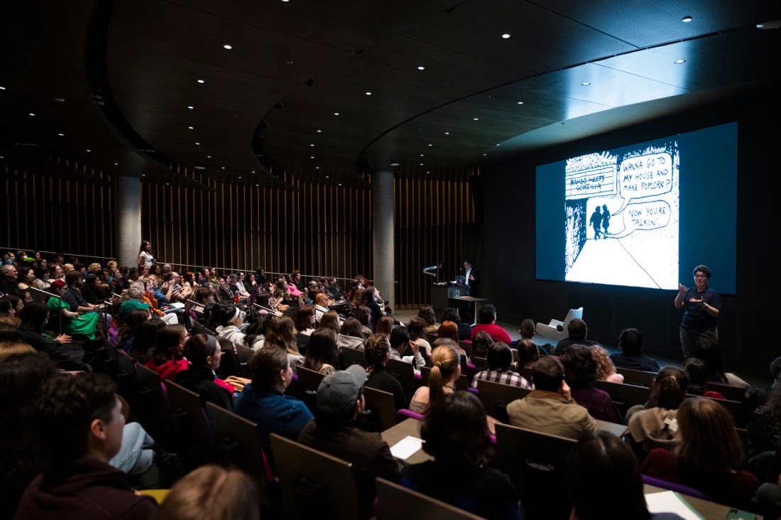 Alison Bechdel standing on stage next to a projector screen showing a comic panel.