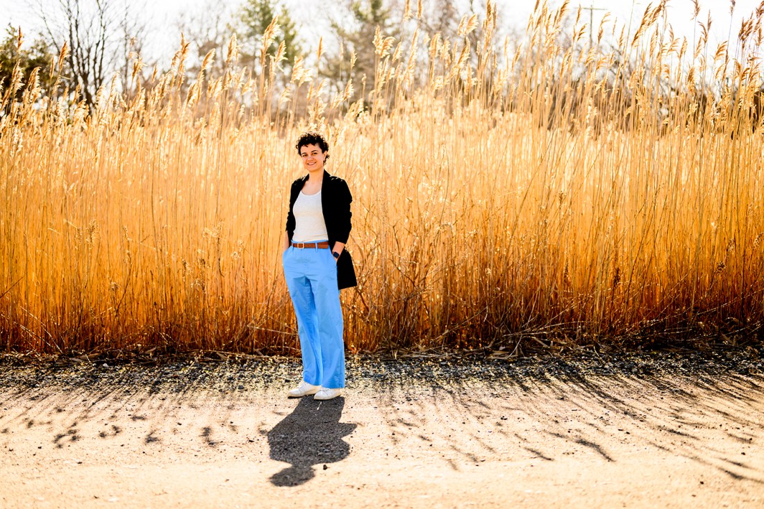 Gabriela Garcia, an assistant professor, stands with short brown hair in bright blue pants against a backdrop of tall grass.