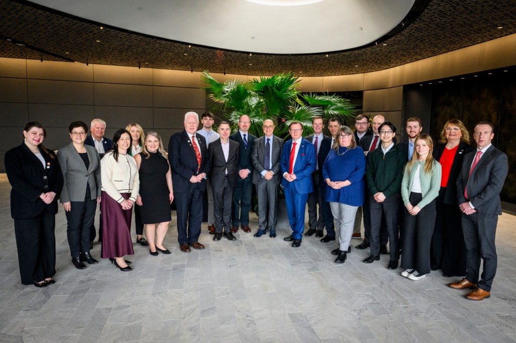 A group of professionally dressed individuals pose for a photo in a modern indoor setting with a plant arrangement in the background.