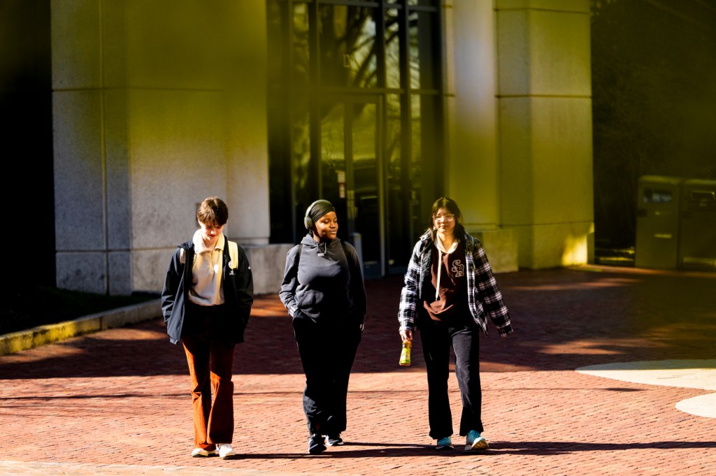 Three people walk together outside on a sunny day, with warm light casting shadows on the brick pathway.