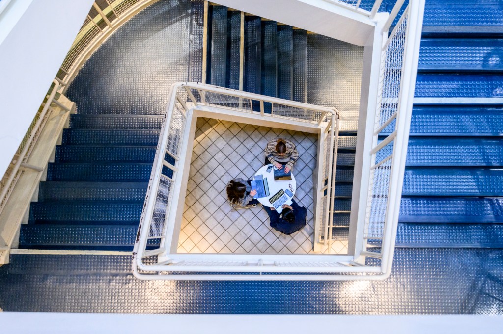 A group of people sit around a table working together, viewed from above through a staircase with blue steps.