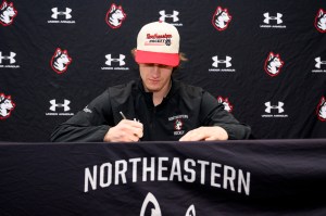 A person wearing sports apparel and a hat sits at a table, signing a document in front of a branded backdrop.