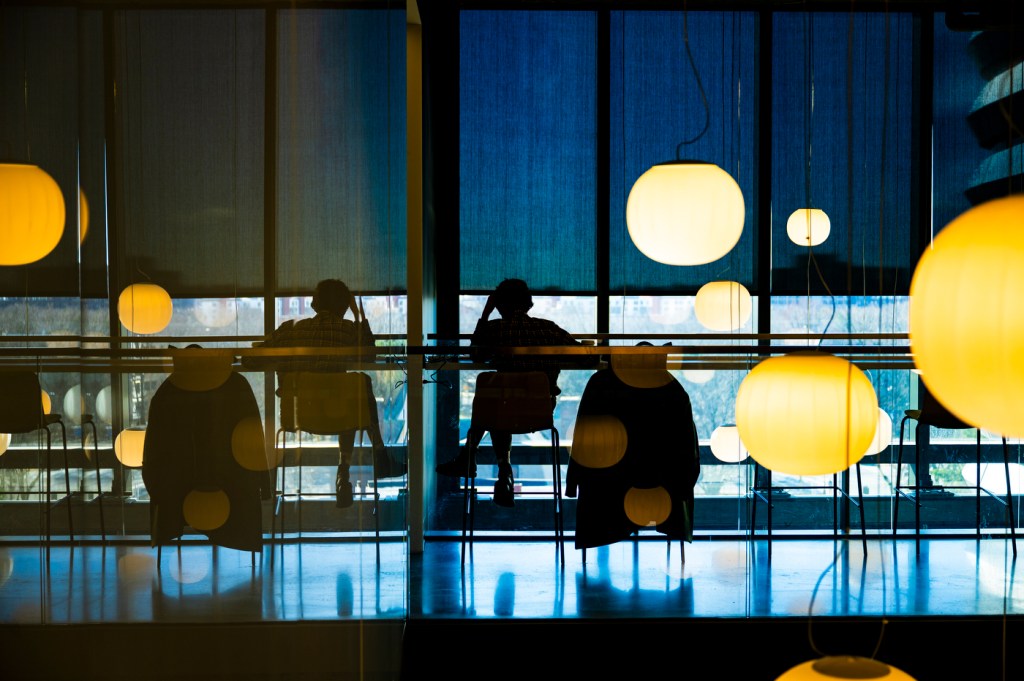 Silhouetted figures sit in a modern interior space, surrounded by hanging lights and large windows with reflections and shadows, creating a layered effect.