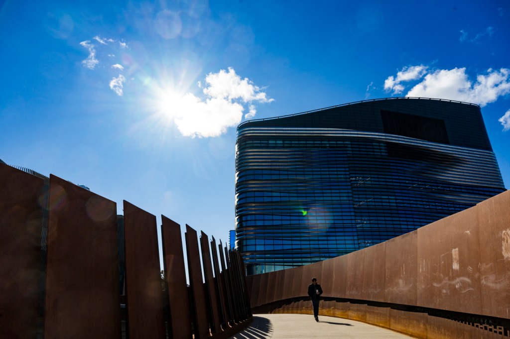 A person walks along a modern pedestrian bridge beneath a bright sky, with a contemporary building visible in the background.