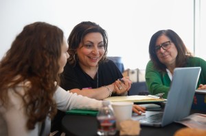 Students sitting next to a small business owner in front of a laptop, teaching them how to use AI.