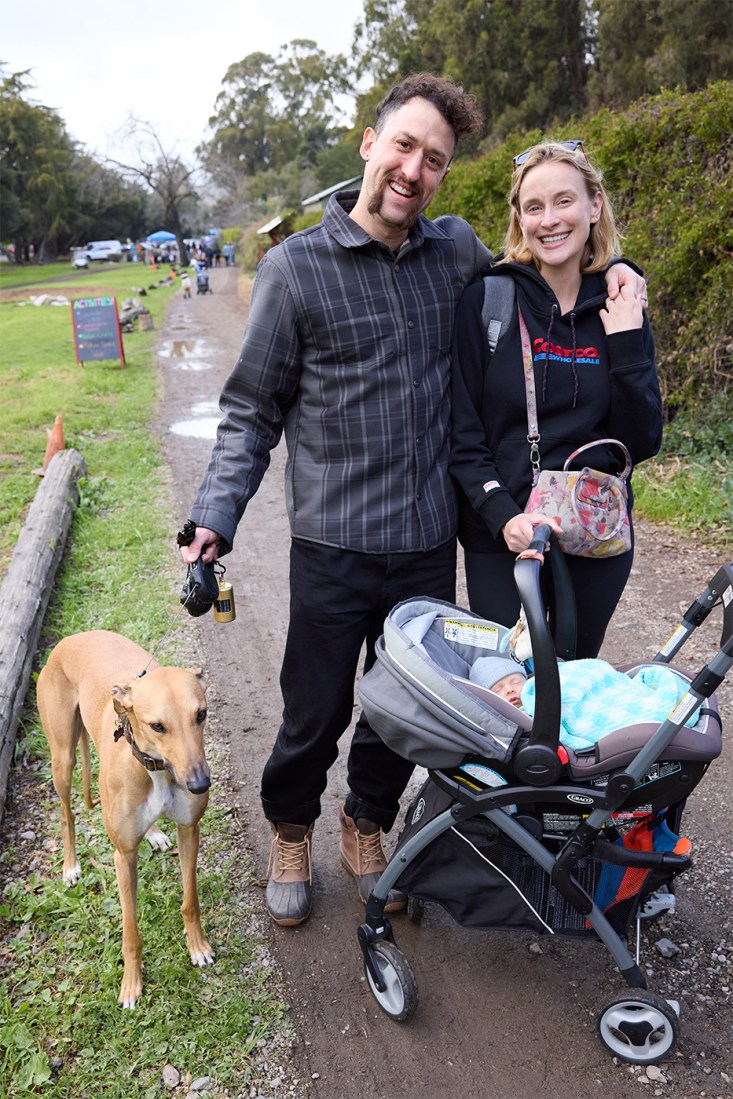 Two people with a dog on a leash and a baby in a stroller standing with their arms around each other. 