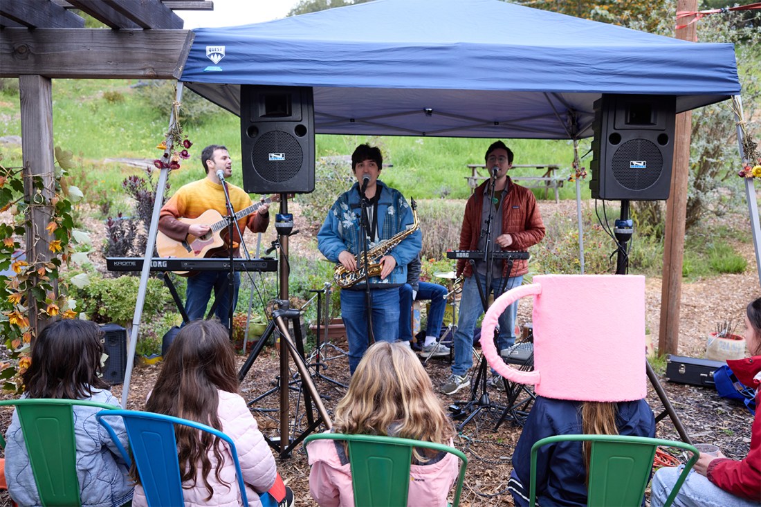 Three musicians playing guitars and singing on an elevated stage underneath a blue tent.