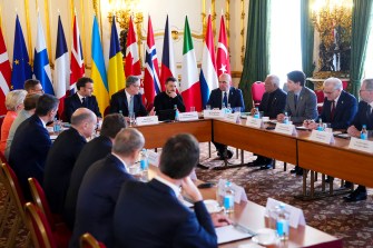 World leaders sitting around a square shaped table at a summit. Next to the table are a series of flags representing the countries that the leaders represent.