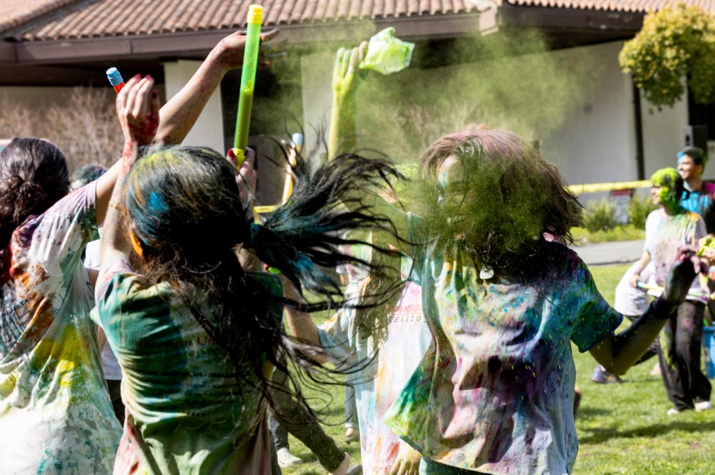 People gather outdoors, throwing colorful powder into the air during a festive celebration.