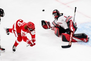 Two hockey players on opposing teams. One is wearing a red jersey and taking a shot on net. The other is a goalkeeper wearing a white jersey making a save.