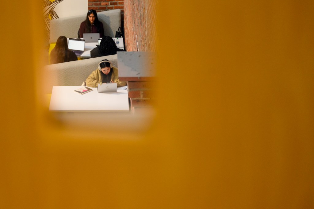 Several people are seen working on laptops in a shared indoor space, viewed through a narrow opening or window in the foreground.