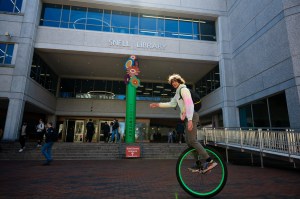 A person rides a unicycle in front of a large building entrance, with several other people walking nearby on a sunny day.