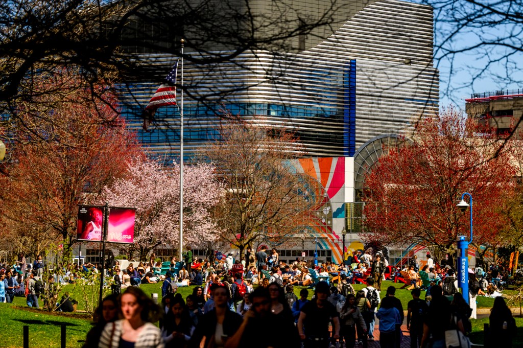 A large group of people gather and walk through a green outdoor space on a sunny day, with blooming trees and a modern building in the background.