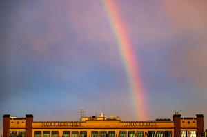 A colorful rainbow arcs across the sky above the top of a building at sunset, with soft lighting and pastel tones in the background.