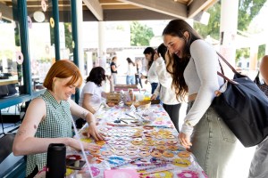 Two people smile and interact at a colorful table covered with round treats and decorations, while others gather in the background.