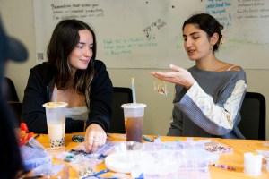 Two people sit at a table working with colorful beads and materials, with drinks nearby and writing visible on a whiteboard in the background.