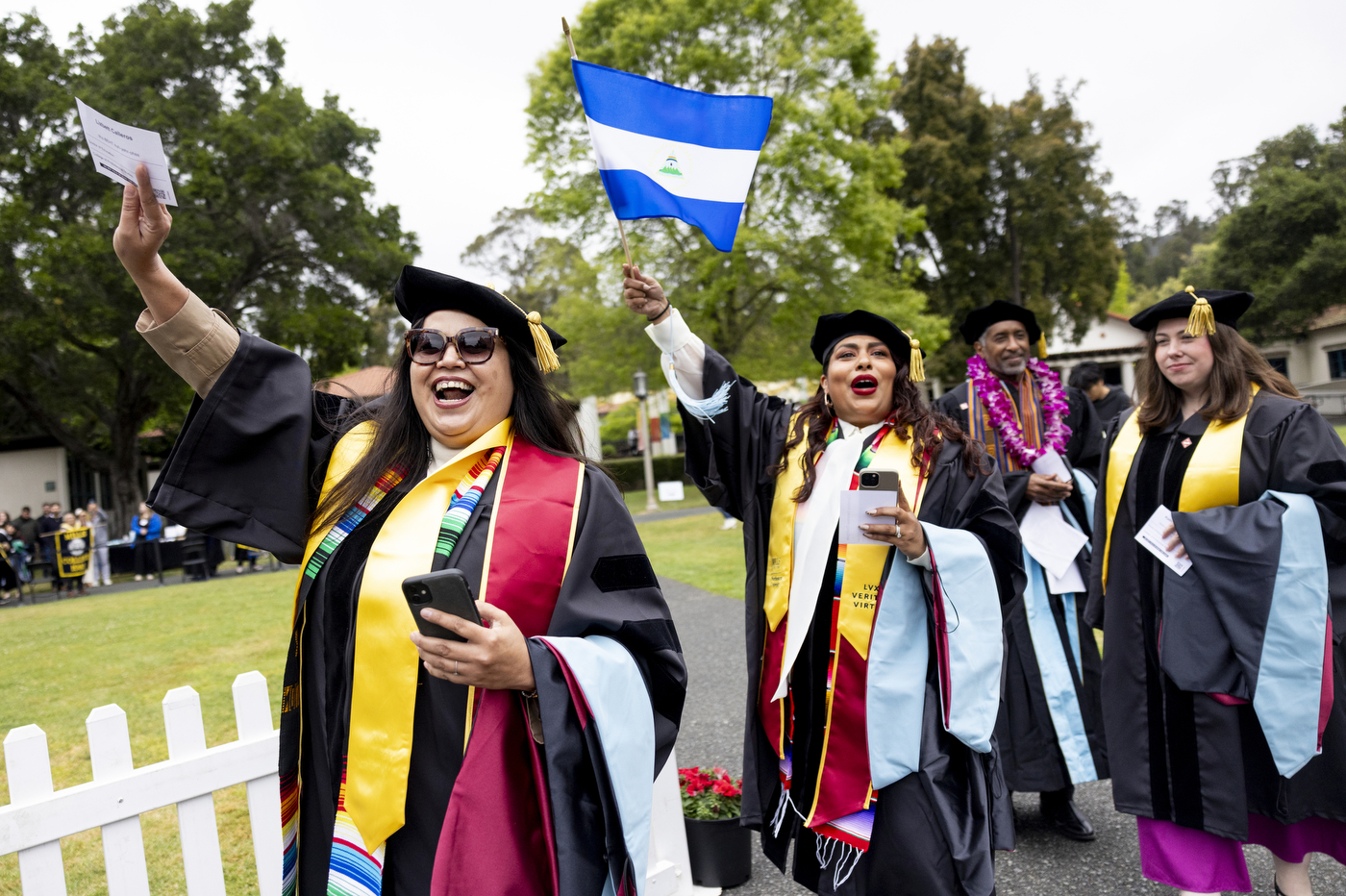 Graduates in academic regalia wave international flags and celebrate outdoors on a lawn with a white picket fence and trees in the background.