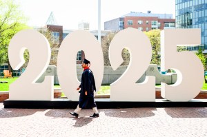 A person wearing graduation attire walks past large numbers displayed outdoors, with buildings and greenery in the background.