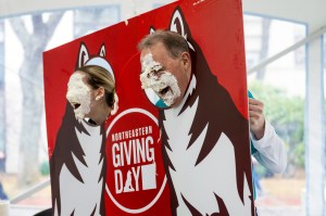 Two members of the Northeastern community with pie covering their faces posing in a Husky photo background.