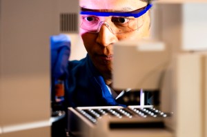 Jing-Ke Weng wearing safety glasses and blue gloves looking intently at a sample in the lab.