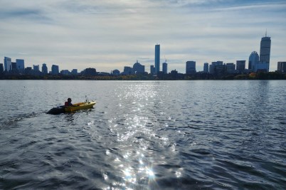 A person kayaks in the Charles River with the Boston skyline visible on the horizon.