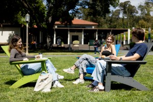 Three people sit in colorful outdoor chairs on a grassy lawn, enjoying a sunny day and chatting with drinks and food in hand.