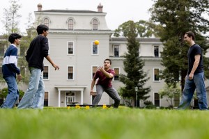 Four people play a ball game on a grassy field in front of a large building, with trees and overcast skies in the background.
