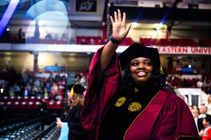 A person in graduation attire smiles and waves while walking through an indoor arena filled with people.