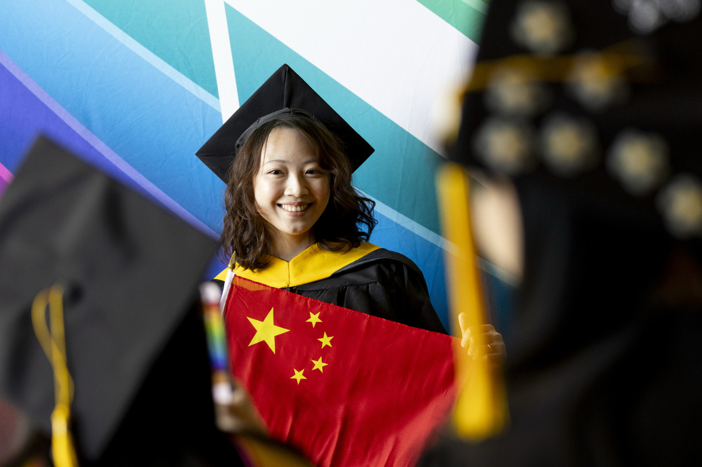 A smiling graduate in a cap and gown holds a Chinese flag and poses for a photo at commencement. 